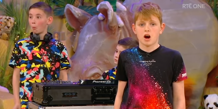 Two little boys behind a set of DJ decks on the toy show set, with one boy in front singing