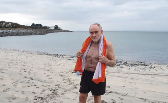 Man standing on a beach in swimming trunks with an orange and white towel wrapped around him, looking at the camera