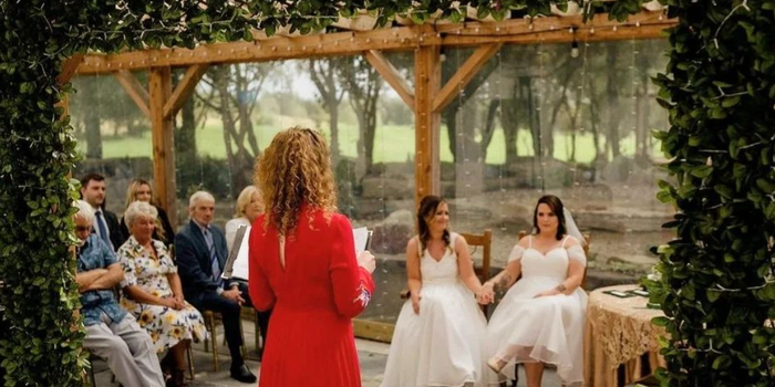 scene from an outdoor wedding with greenery and wooden beams in the background - two brides holding hands, and an officiator reading from a book
