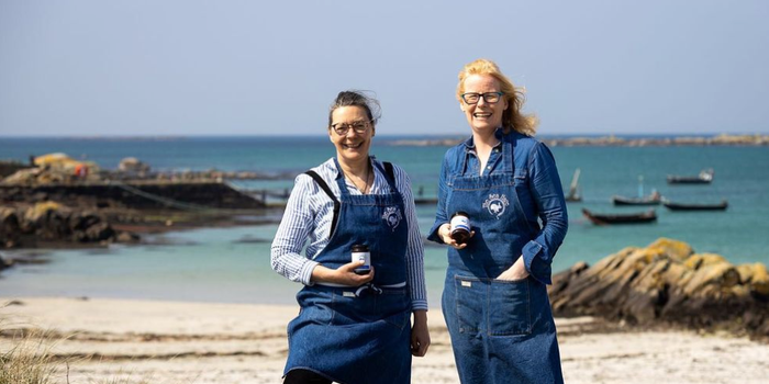 the two owners of the Sea Hare with aprons and coffee cups smiling with a beach in the background