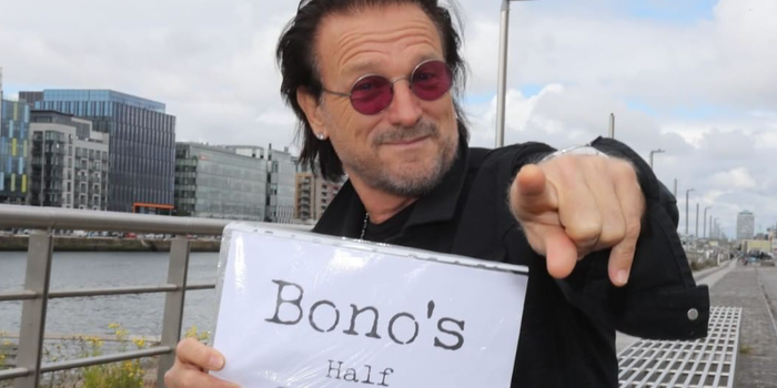 Bono lookalike sitting on a bench by a river holding a sign that reads "Bono's half brother"