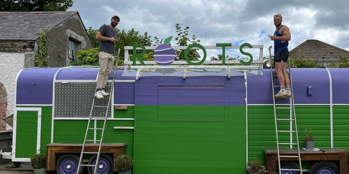 large horsebox cafe painted purple and green, with a sign that reads "roots". two men on ladders working on the horsebox, posing for camera