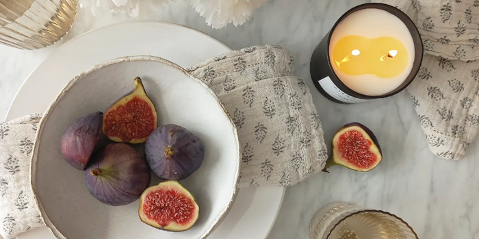 flat lay of a lit candle on a table with white tablecloth, matching napkins and a bowl of figs.