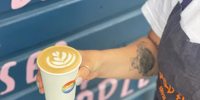 person holding a milky coffee with latte art, wall of a horsebox cafe can be seen in the background