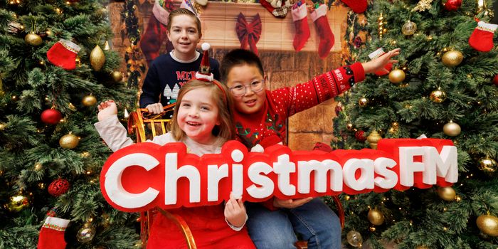 children in front of a fireplace decorated for christmas and christmas trees on either side, holding a sign that says Christmas FM